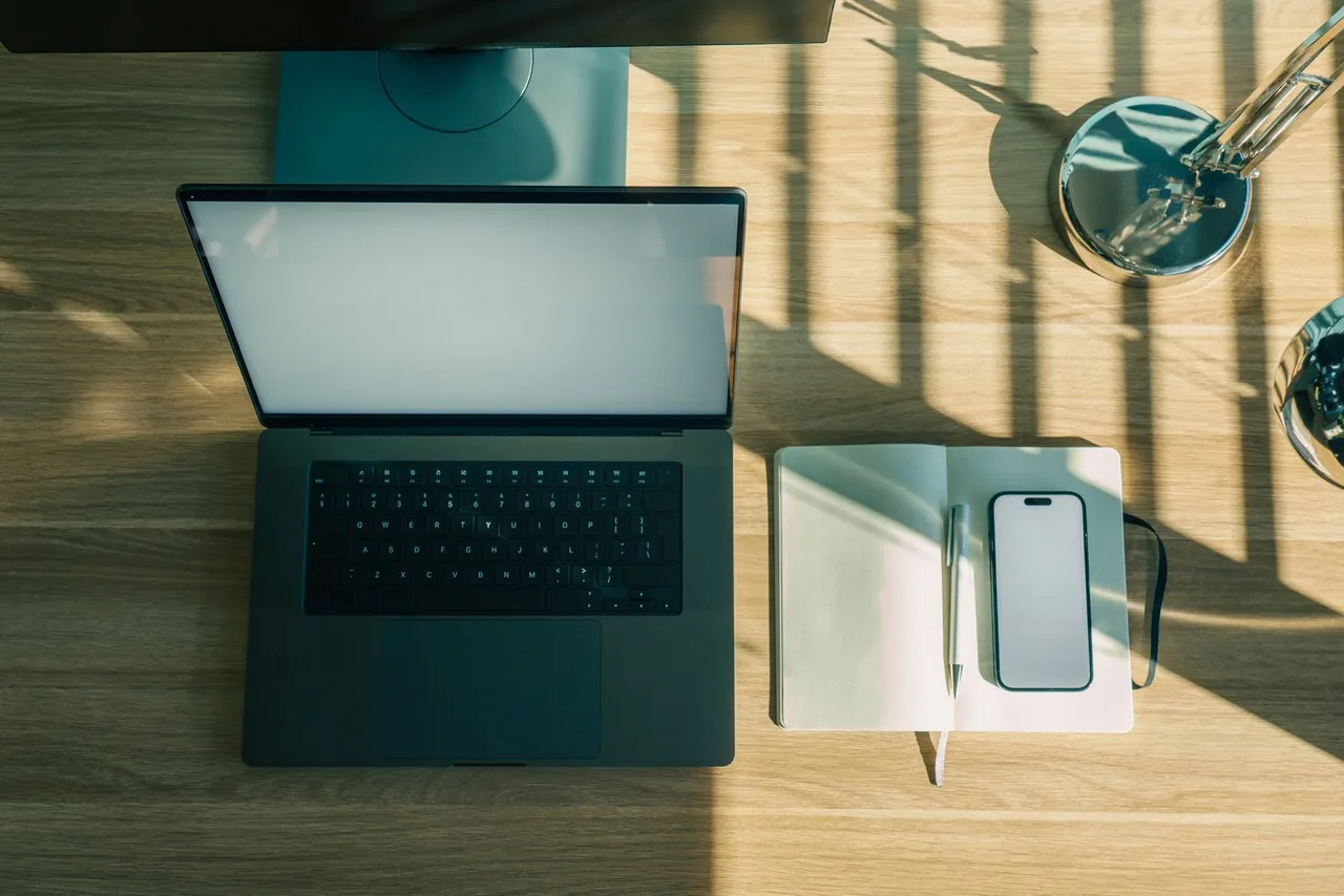 An overhead view of a laptop beside an open notebook, pen, and smartphone on a sunlit wooden desk.